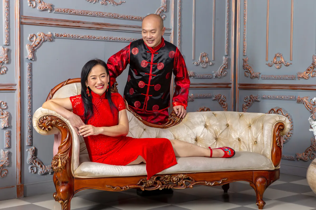 ธนินพงษ์ ศุภพิทักษ์พงษ์ ในชุดจีนสีดำแดง ยืนพิงพนักโซฟาข้างภรรยาที่นั่งในชุดกี่เพ้าสีแดง