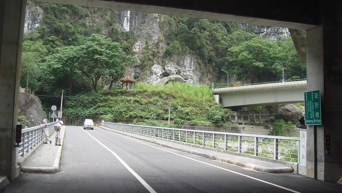 สะพานจิ้นเหิง 靳珩橋 ในช่องเขาทาโรโกะ ไต้หวัน มองจากใต้อุโมงค์ ถนนข้ามสะพานราวเหล็กสีขาว หน้าผาหินปูน และป้ายเขียวชื่อสะพาน ตั้งชื่อรำลึกวิศวกรแซ่จิ้น