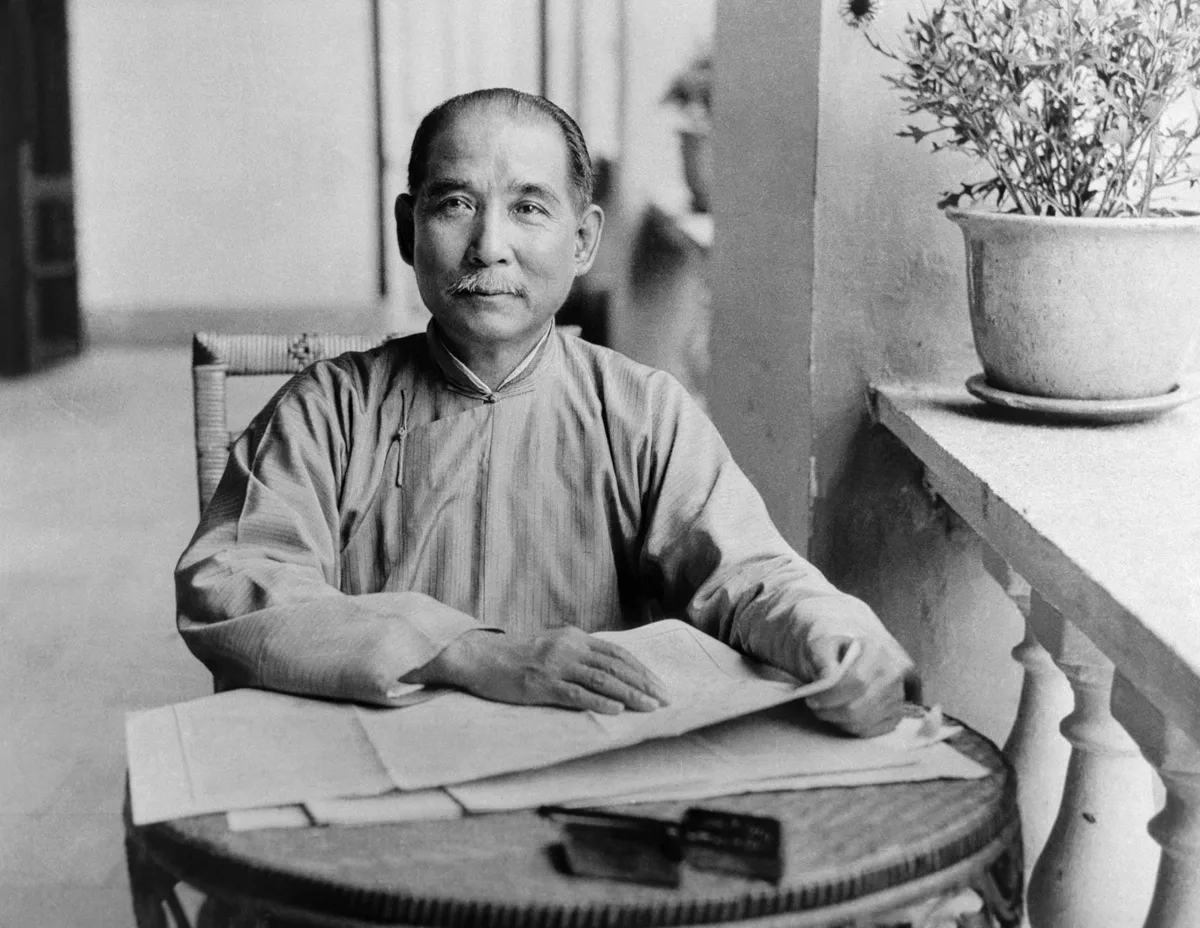 ภาพขาวดำ ดร.ซุนยัตเซ็น คนแซ่ซึง แซ่ซุน นั่งที่โต๊ะไม้กลม มีกระดาษวางตรงหน้า ข้างกระถางต้นไม้ริมระเบียง