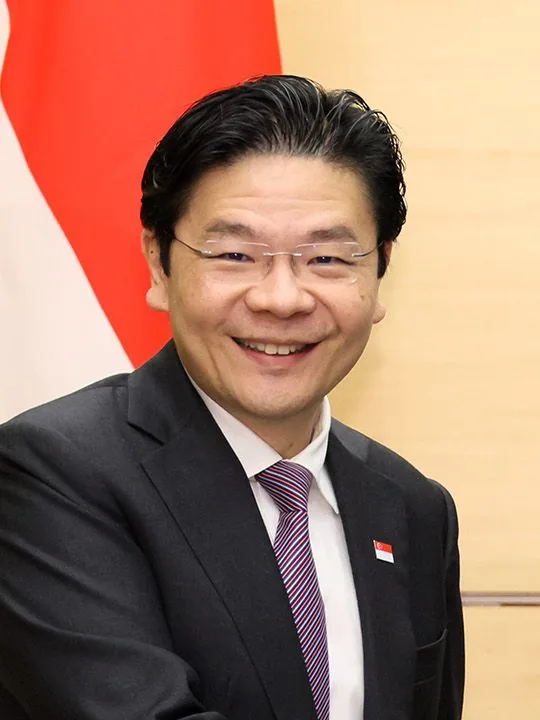 ลอว์เรนซ์ หว่อง ในชุดสูท ติดเข็มกลัดธงชาติสิงคโปร์ ยืนหน้าธงชาติ