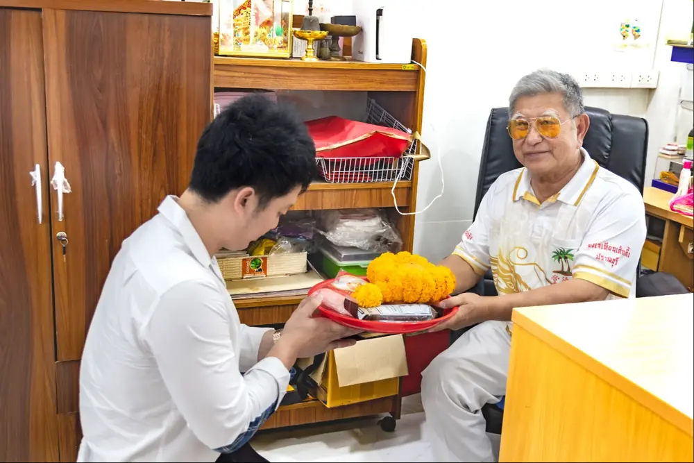 ปัญญวัฒน์ ศุภพิทักษ์พงษ์ (Ken) ในเสื้อเชิ้ตขาว ยกถาดไหว้ครูที่มีพวงดาวเรืองสีส้มให้ซินแสตี๋ผู้นั่งบนเก้าอี้