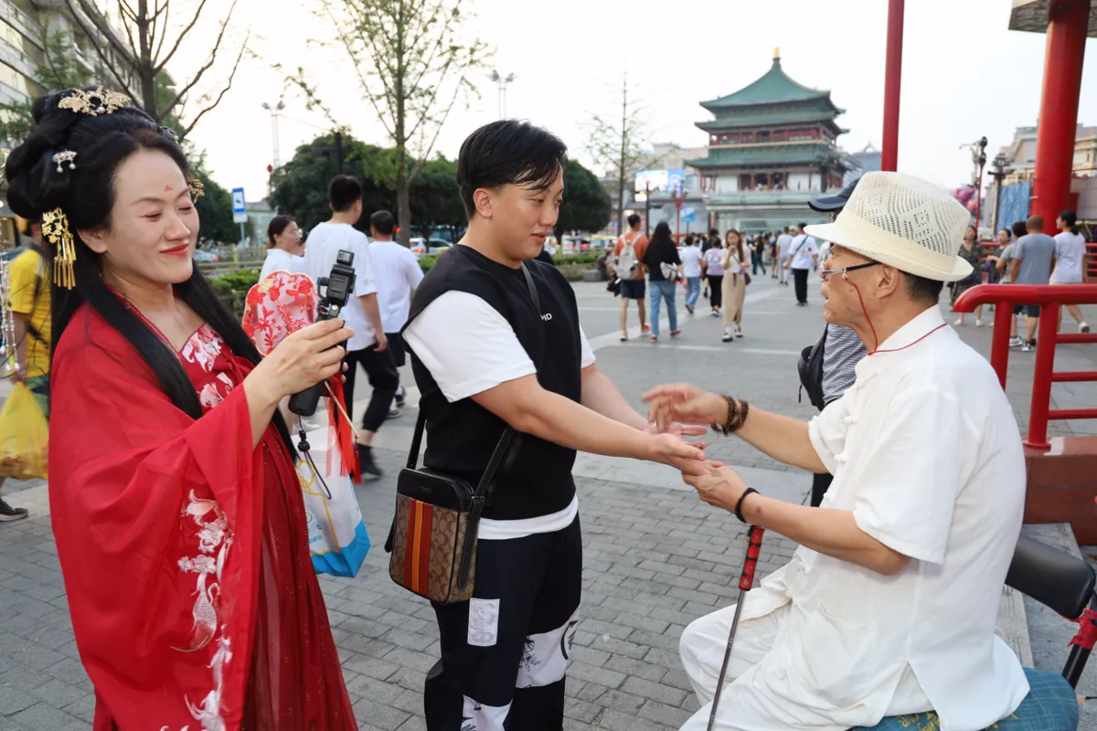 ซินแสริมทางในชุดขาวสวมหมวกสานจับมือชายหนุ่มดูลายมือ ขณะหญิงชุดฮั่นฝูสีแดงถ่ายวิดีโอ มีหอระฆังซีอานเป็นฉากหลัง